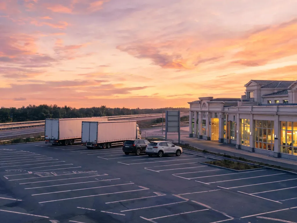 Nowoczesny parking Autohof o zachodzie słońca z ciężarówkami, autami i oświetlonym budynkiem usługowym przy autostradzie.